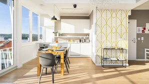 Eine moderne Küche mit weißen Schränken, eine Essecke mit Holztisch und Stühlen sowie ein Blick auf die Stadt durch große Fenster.