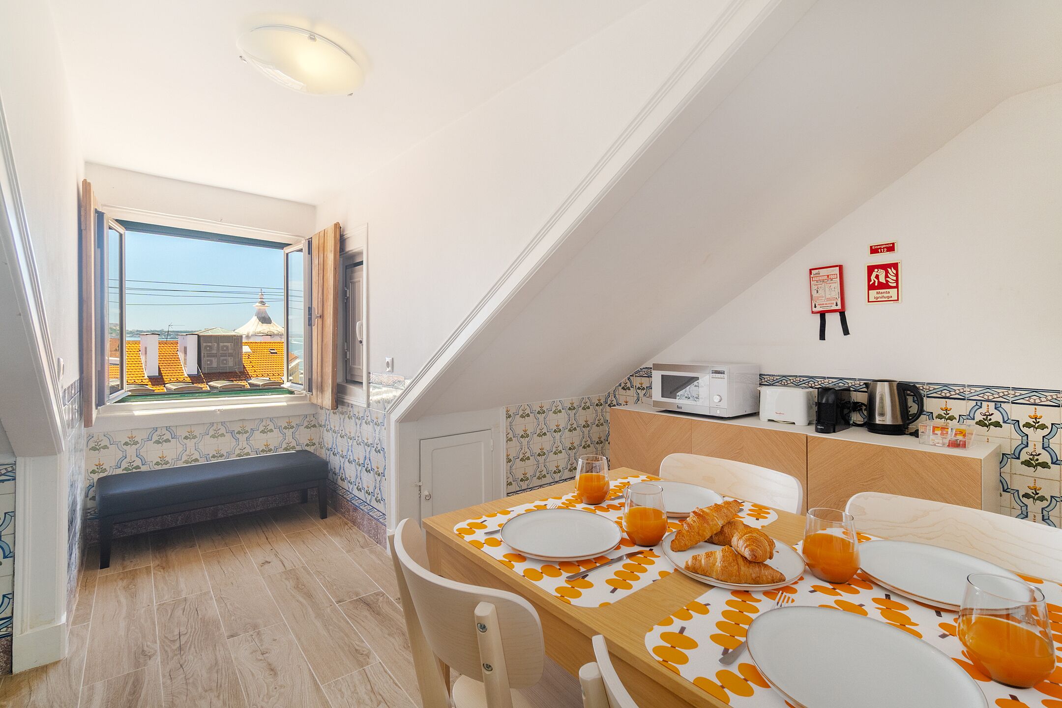 Un coin repas avec une table mise pour le petit-déjeuner, un banc et une vue sur un bâtiment par une fenêtre ouverte.