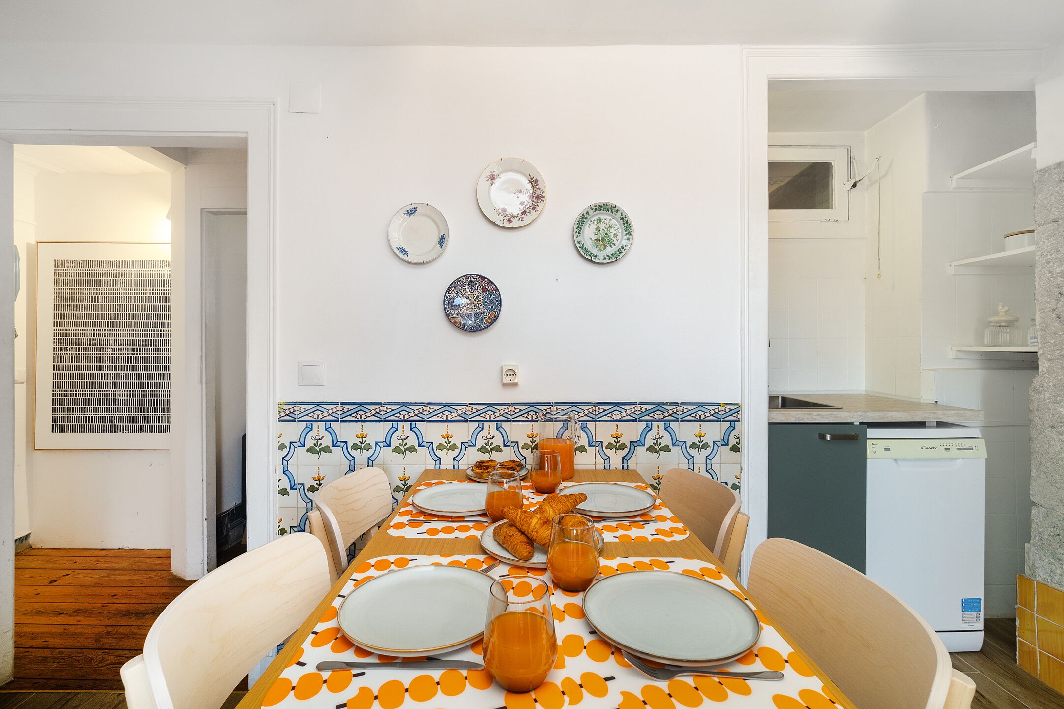 Un comedor con una mesa dispuesta para cuatro personas, rodeada de sillas, y una zona de cocina con lavavajillas y armarios.