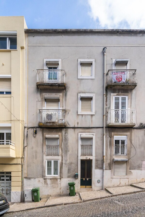 Un edificio con varios balcones, una puerta con un número y dos contenedores verdes para basura.