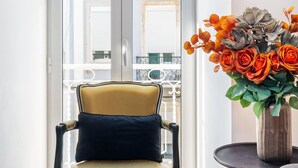 Une pièce avec un plancher en bois, une chaise jaune avec un coussin noir, un fauteuil gris, une petite table ronde ornée d’un vase de fleurs et une porte donnant sur un balcon avec une balustrade.