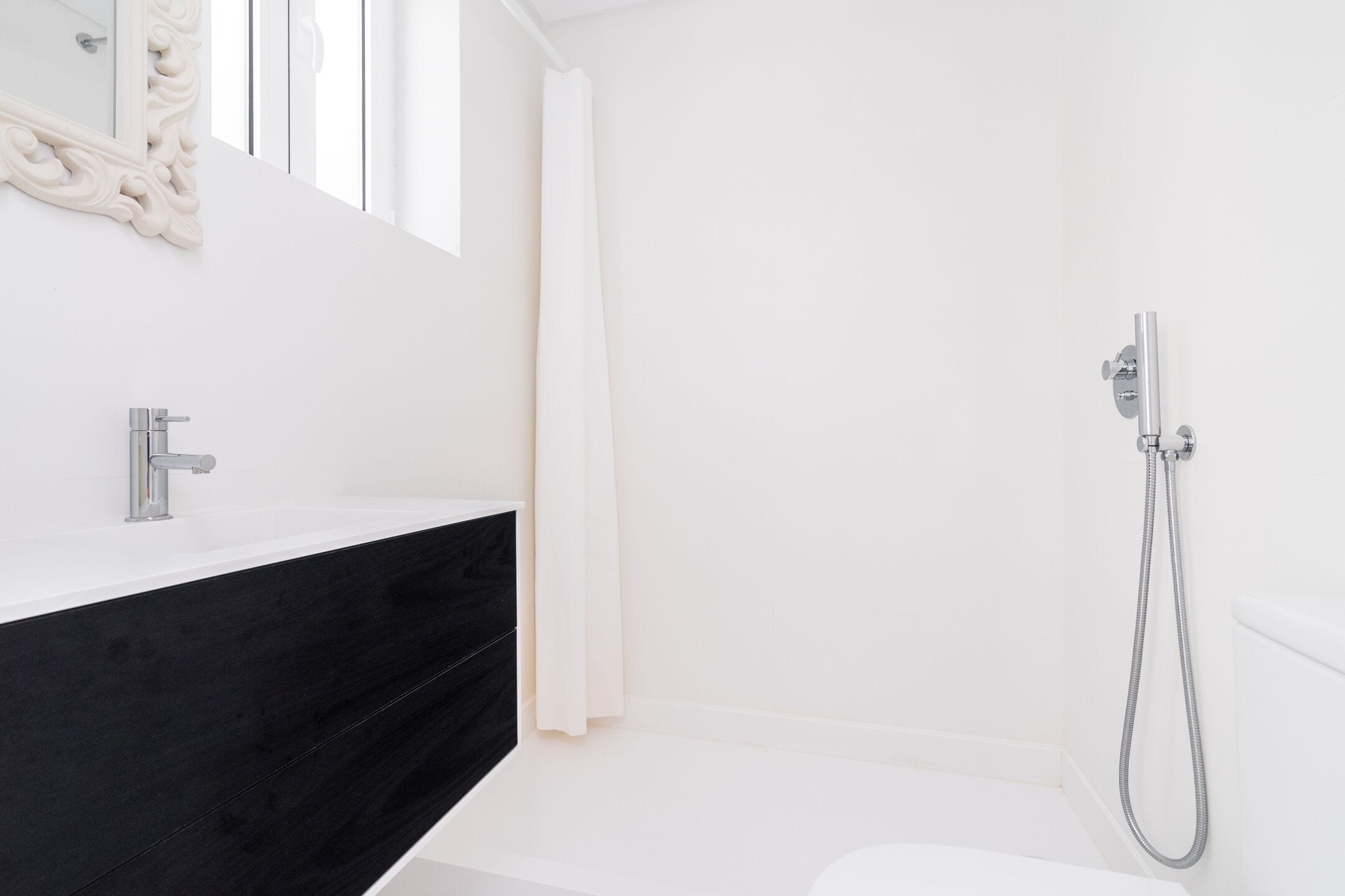 Un baño moderno con tocador blanco, encimera de mármol negro, ducha con cabezal de ducha de mano y un espejo grande con detalles ornamentales.