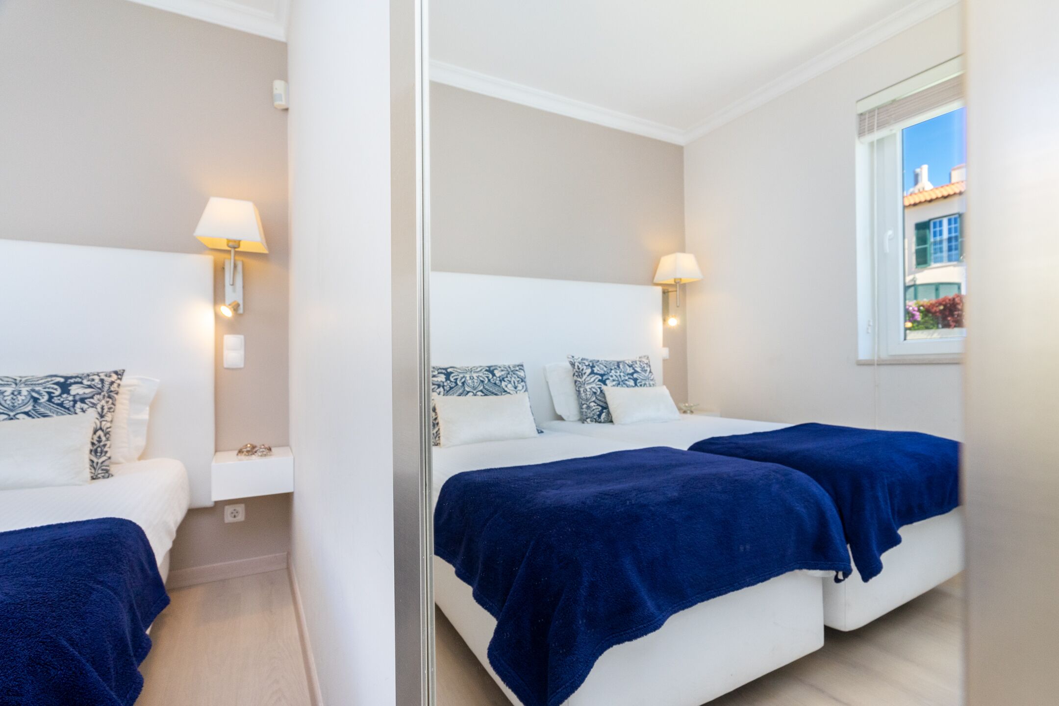 A hotel room with two beds, each with a blue blanket, white bedding, and patterned pillows. There are wall-mounted lamps on either side of the beds.