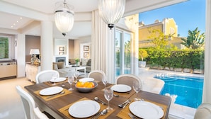 Un comedor con una mesa dispuesta para seis personas, una piscina visible a través de la ventana y una cocina al fondo.
