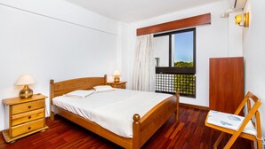 A neatly arranged bedroom with a wooden bed, bedside tables with lamps, a wooden chair, and a window with a view of greenery.