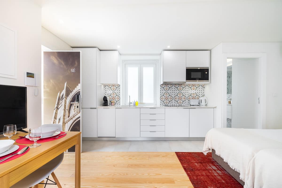 Ein modernes Hotelzimmer mit einem Holzespeisetisch, gedeckt für zwei Personen, einem Bett mit rotem Teppich, einer Küche mit weißen Schränken und einem großen Fenster.
