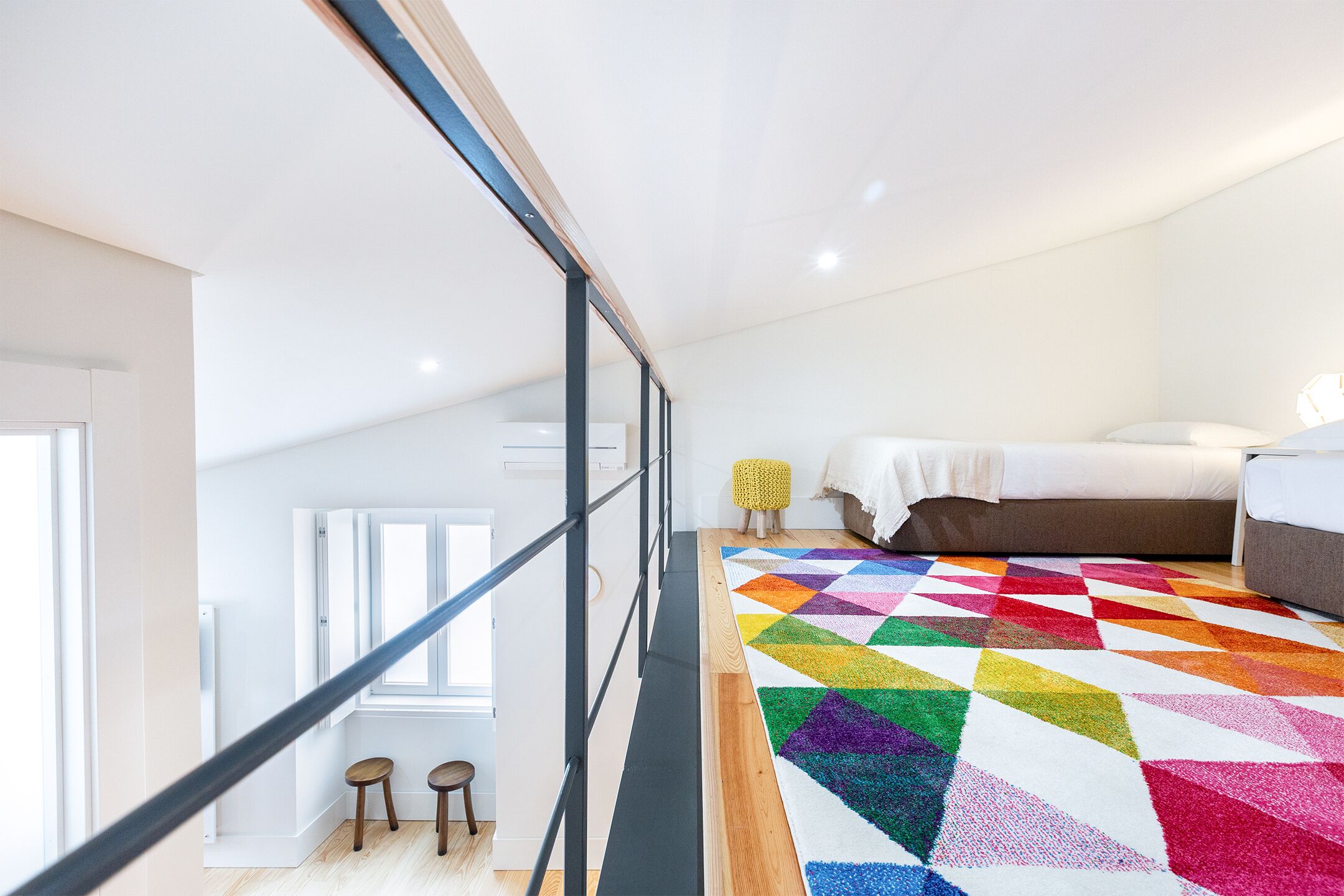 A modern bedroom with a colorful geometric rug, a bed, and a staircase leading to an upper level.
