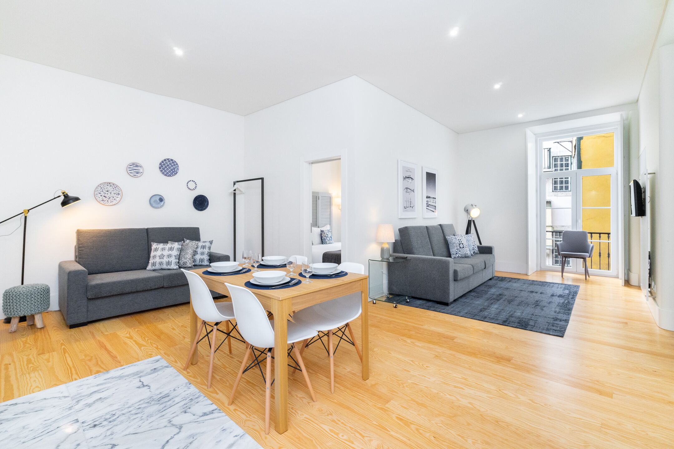 A modern living room with a dining area, grey sofas, a wooden dining table, and a large window.