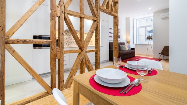 Uma área de refeições com uma mesa de madeira posta para dois, uma cozinha ao fundo e uma sala de estar com um sofá e um abajur.
