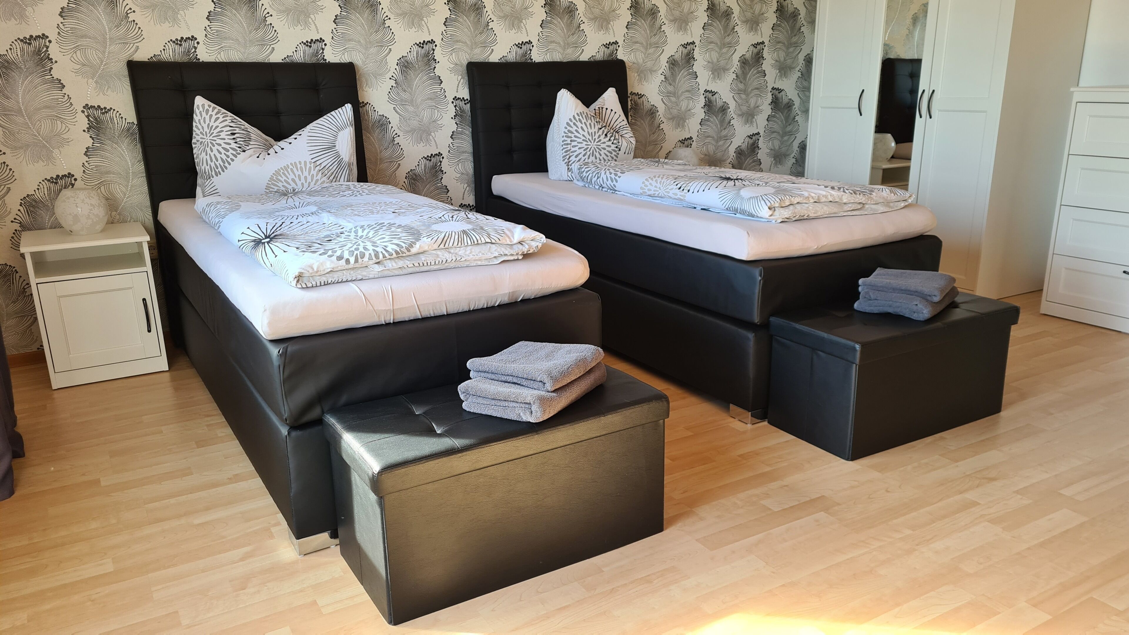 Quarto com duas camas, cada uma com cabeceira preta e roupa de cama branca. Há mesas de cabeceira com abajures, um guarda-roupa e uma pequena mesa de cabeceira com um vaso.