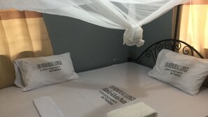 Una cama con sábanas blancas, dos almohadas con el texto 'LM MEMORIAL LODGE' y una toalla blanca doblada, además de un mosquitero.
