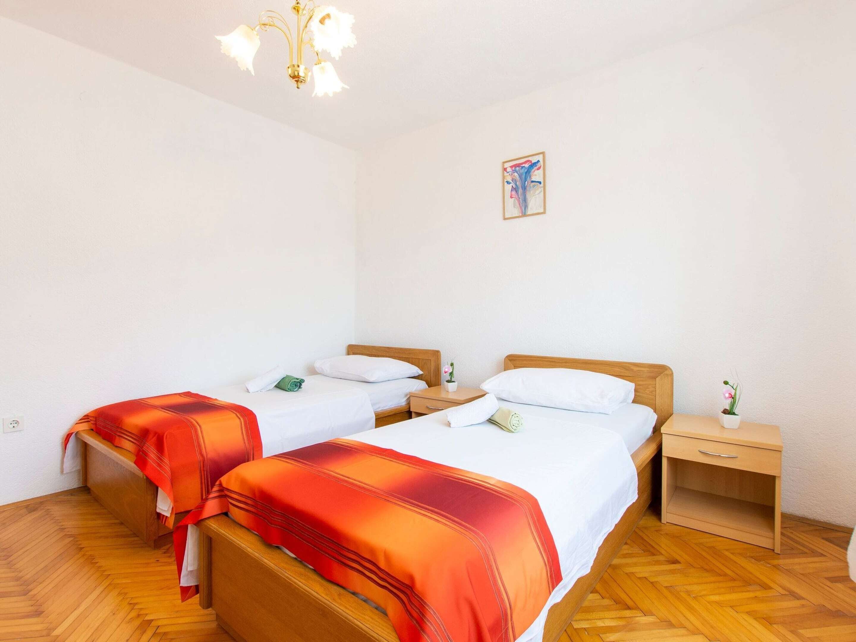A room with two single beds, wooden headboards, a nightstand with a lamp, a small table with a vase, and a painting on the wall.