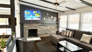 A modern living room with a stone fireplace, a flat-screen TV, a ceiling fan, and a large window with blinds.