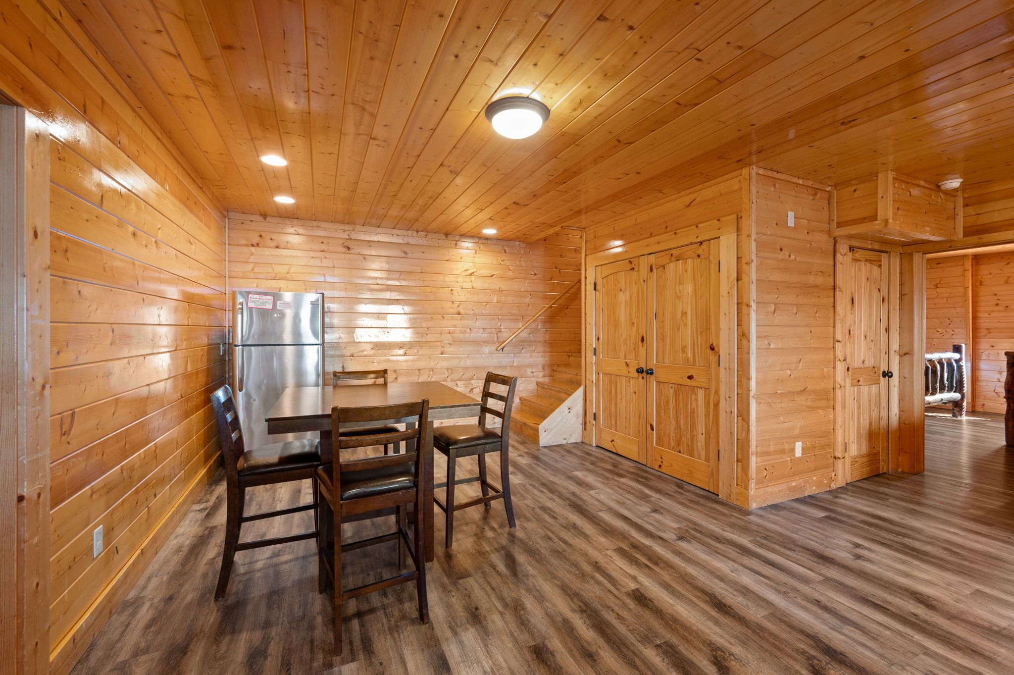 A wooden cabin interior with a dining table, chairs, a refrigerator, and a staircase leading to another room.