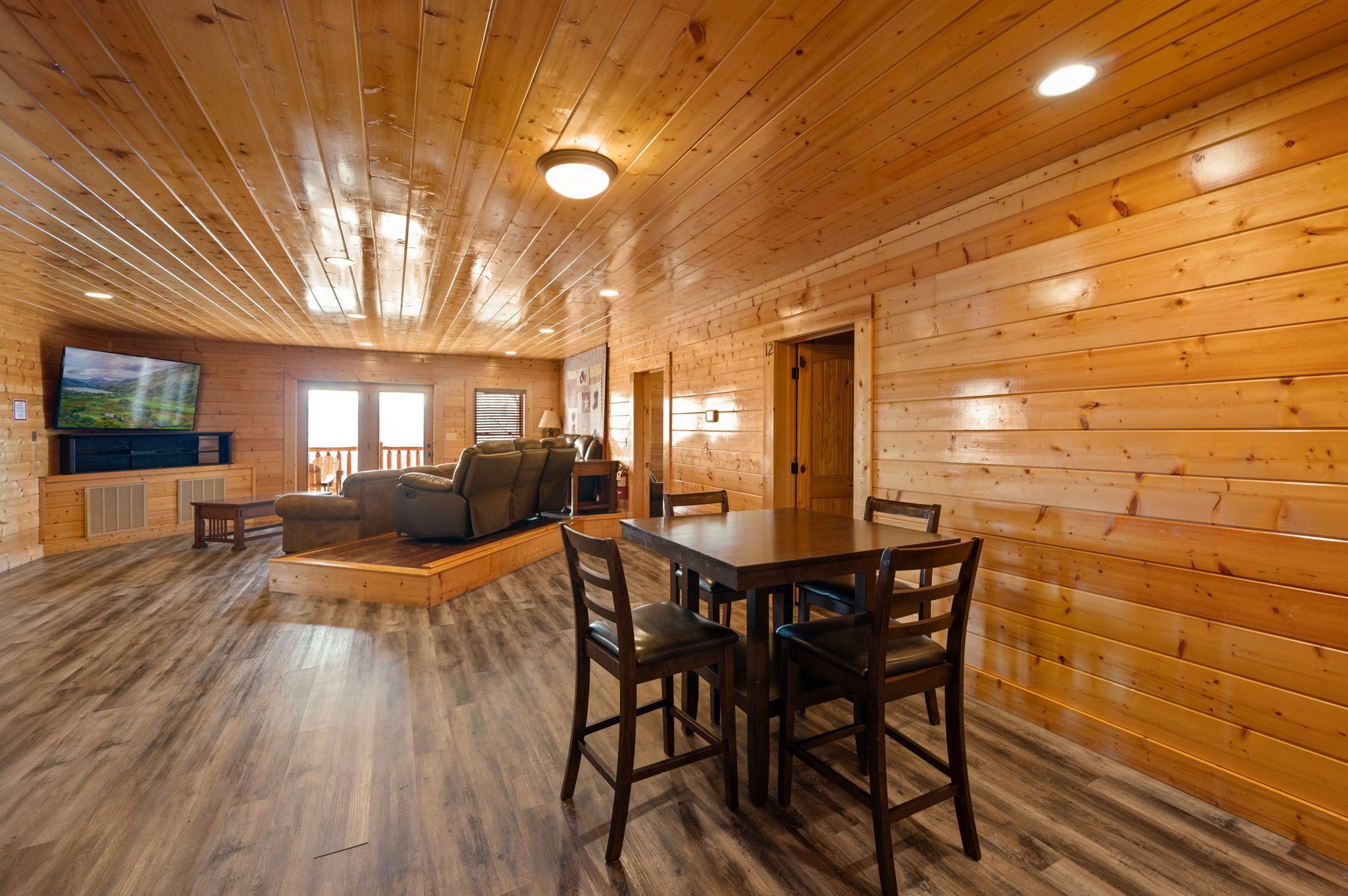 A spacious living room with a wooden floor, a large TV, a sofa, and a dining table with chairs.