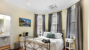 A bedroom with a bed, bedside tables, a nightstand, a window with curtains, a wall-mounted picture, and a bathroom with a sink and mirror. - Cozy Suite in Downtown Charleston with HEATED POOL (Charleston)
