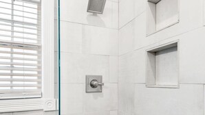 Un baño moderno con azulejos blancos, cabina de ducha de vidrio y ventana con persianas.