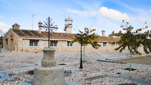 A stone building with a tiled roof, a central stone pillar, and a decorative metal fence. - Complejo Rural Puente Romano (Orgaz)