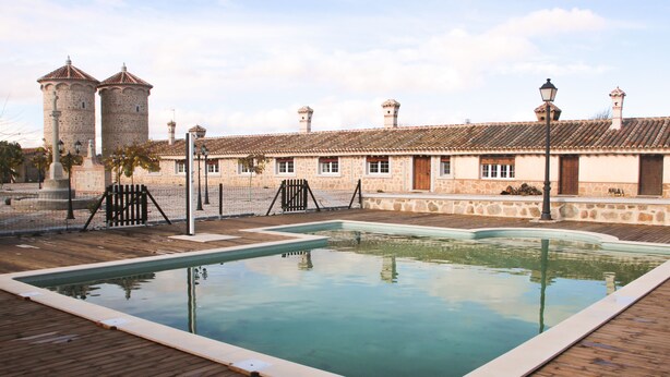 A swimming pool with a wooden deck, stone buildings with tiled roofs, and a fence in the background. - Complejo Rural Puente Romano (Orgaz)