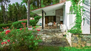 Un patio de piedra que conduce a un área de estar al aire libre techada, con una mesa y sillas, rodeada de exuberante follaje y plantas con flores.