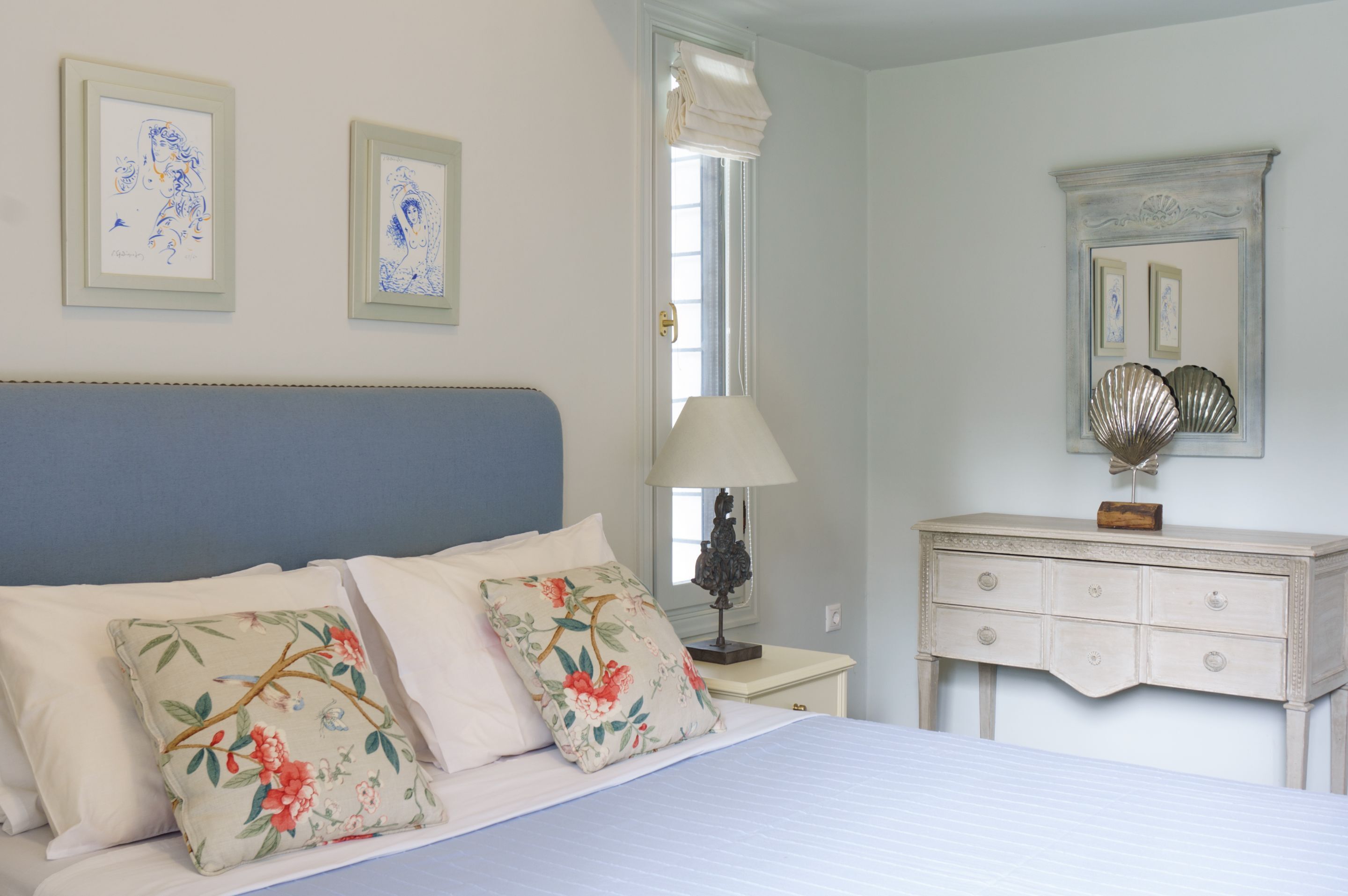 Quarto com cama, travesseiros, mesa de cabeceira, abajur, cômoda e duas obras de arte emolduradas na parede.