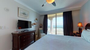 Habitación de hotel con cama, televisor, escritorio, lámpara, silla, ventana con cortinas, ventilador de techo y vista a la playa.