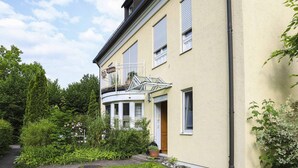 Ein zweistöckiges Gebäude mit einem Balkon, einem Garten mit verschiedenen Pflanzen und einem gepflasterten Weg.