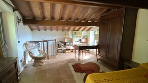Una habitación con techo de madera, un piano, una mesa de comedor con sillas, un sillón de mimbre y una cama con manta amarilla.