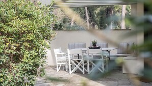 Un patio con mobiliario blanco, una planta en maceta y vistas a árboles y un edificio.