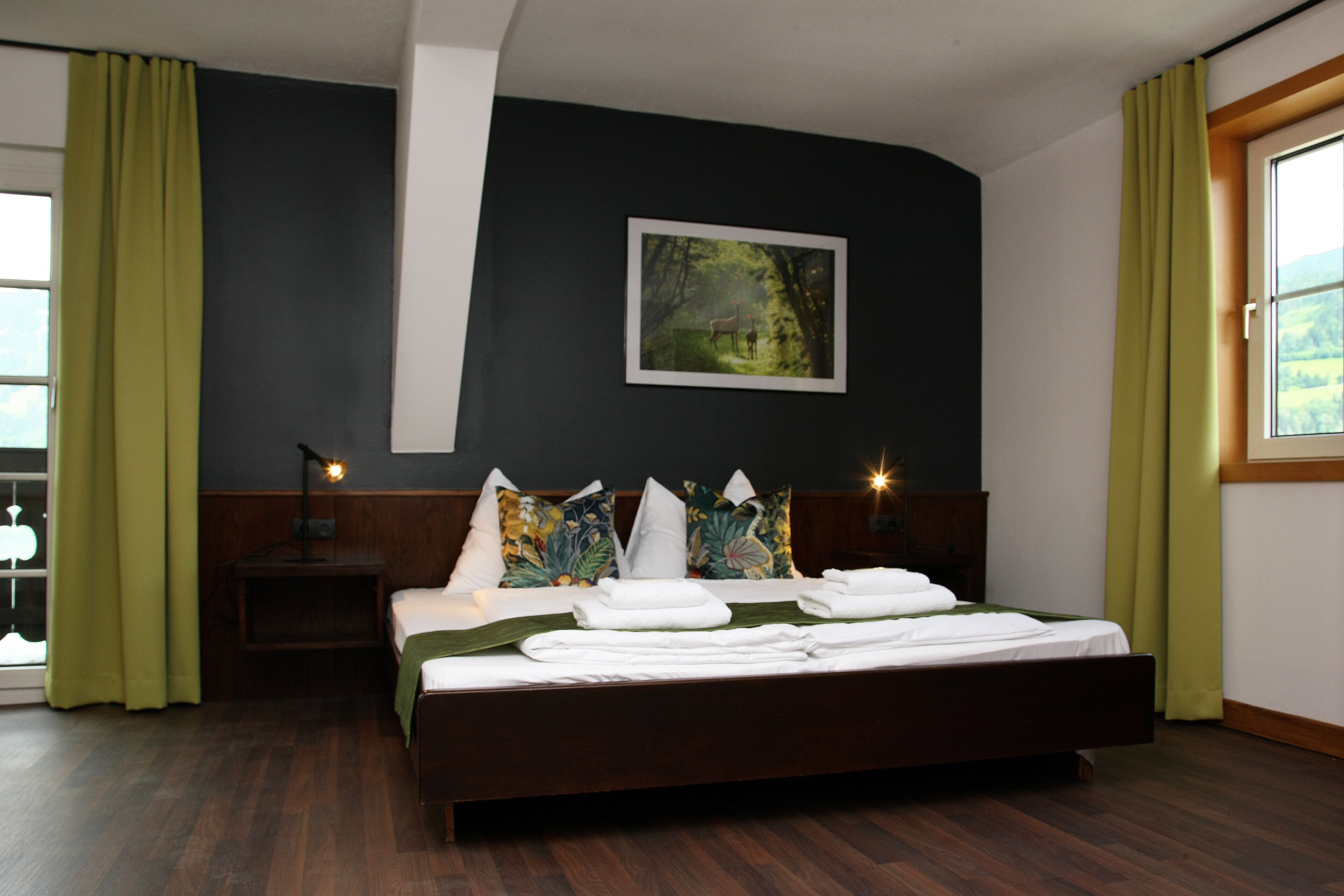 A hotel room with a large bed, green curtains, a nightstand with a lamp, and a painting on the wall.
