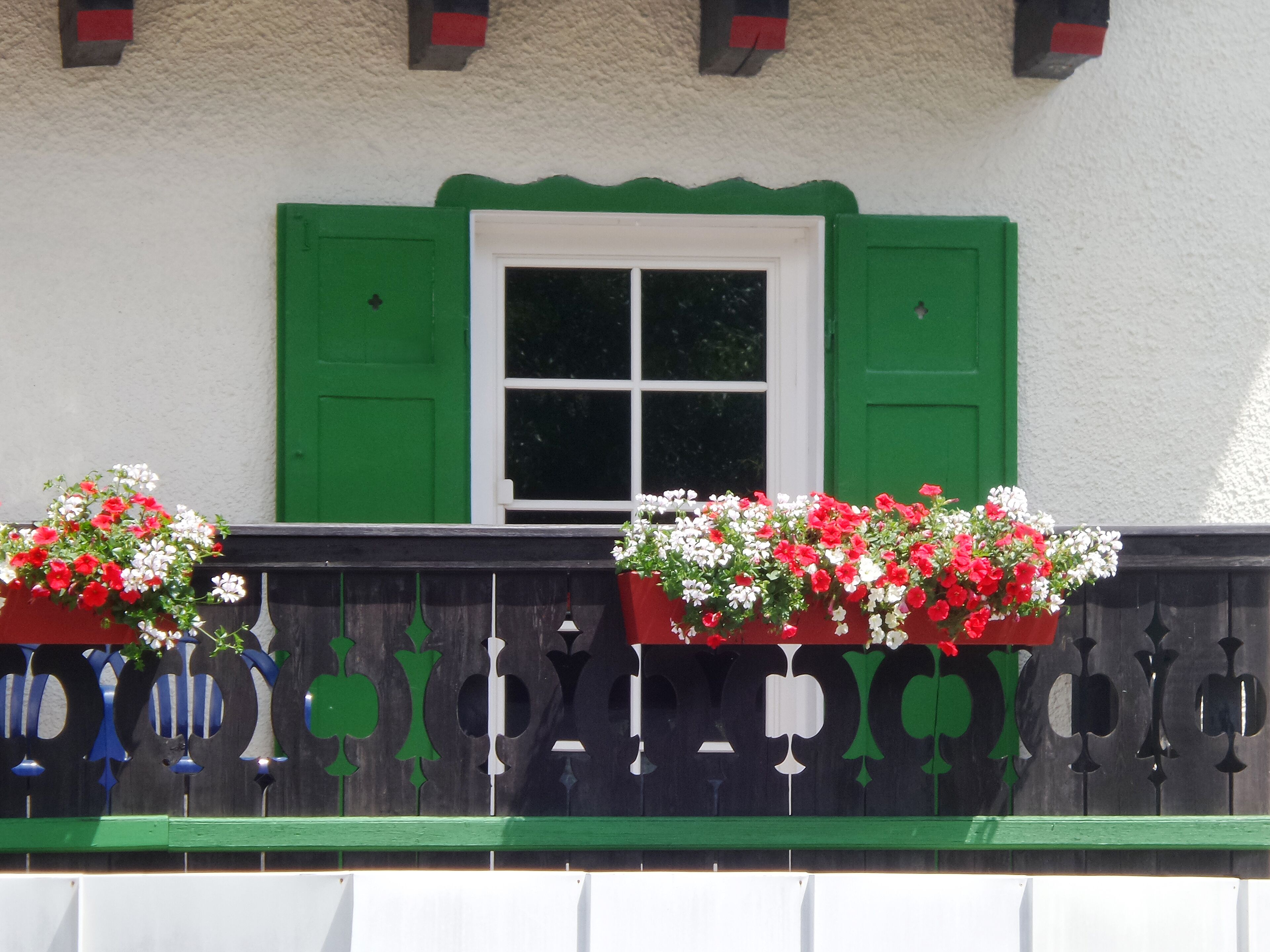 A building with a white exterior, green shutters, and a window with a white frame. There are two flower boxes with red and white flowers on the balcony.