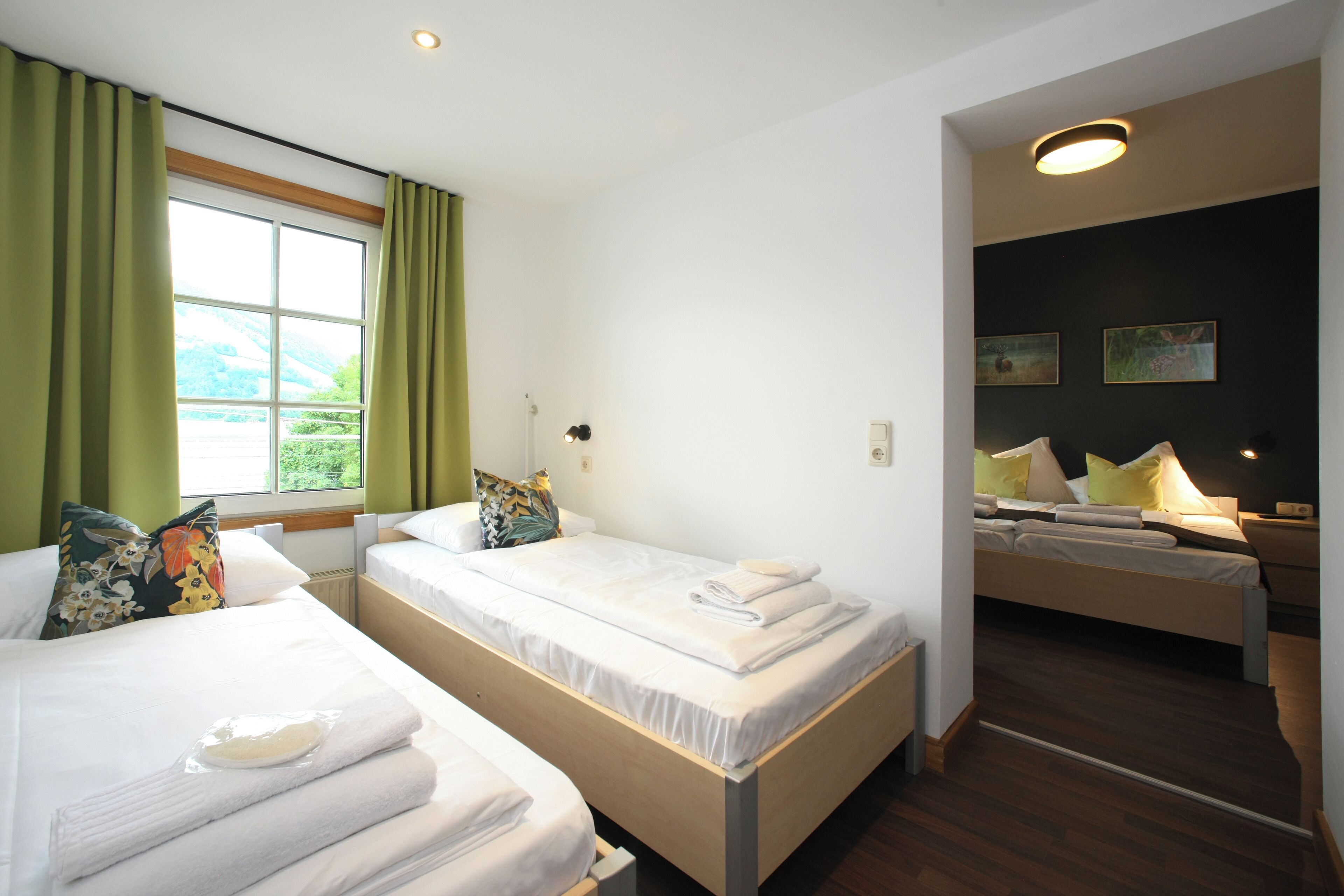 A hotel room with two single beds, a window with curtains, a small table, and a nightstand with a lamp.
