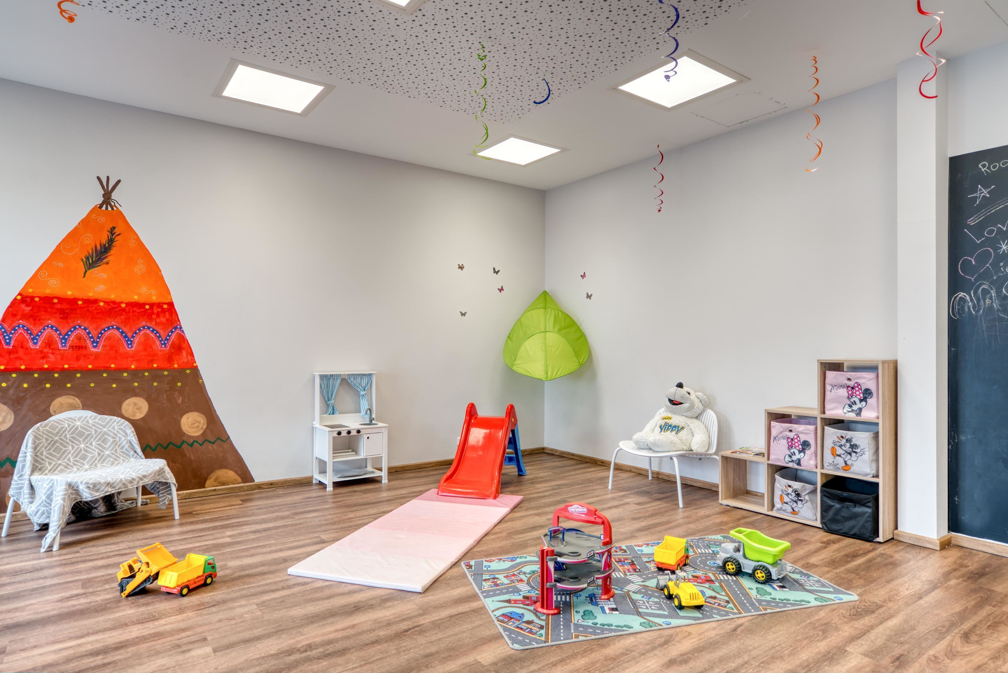 A child-friendly room with a play area, a white chair, a small table, and a chalkboard wall.