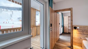 A hotel room with a wooden floor, a large window with a view of a snowy outdoor area, and a wooden door leading to a hallway. - COOEE alpin Hotel Bad Kleinkirchheim (Bad Kleinkirchheim)