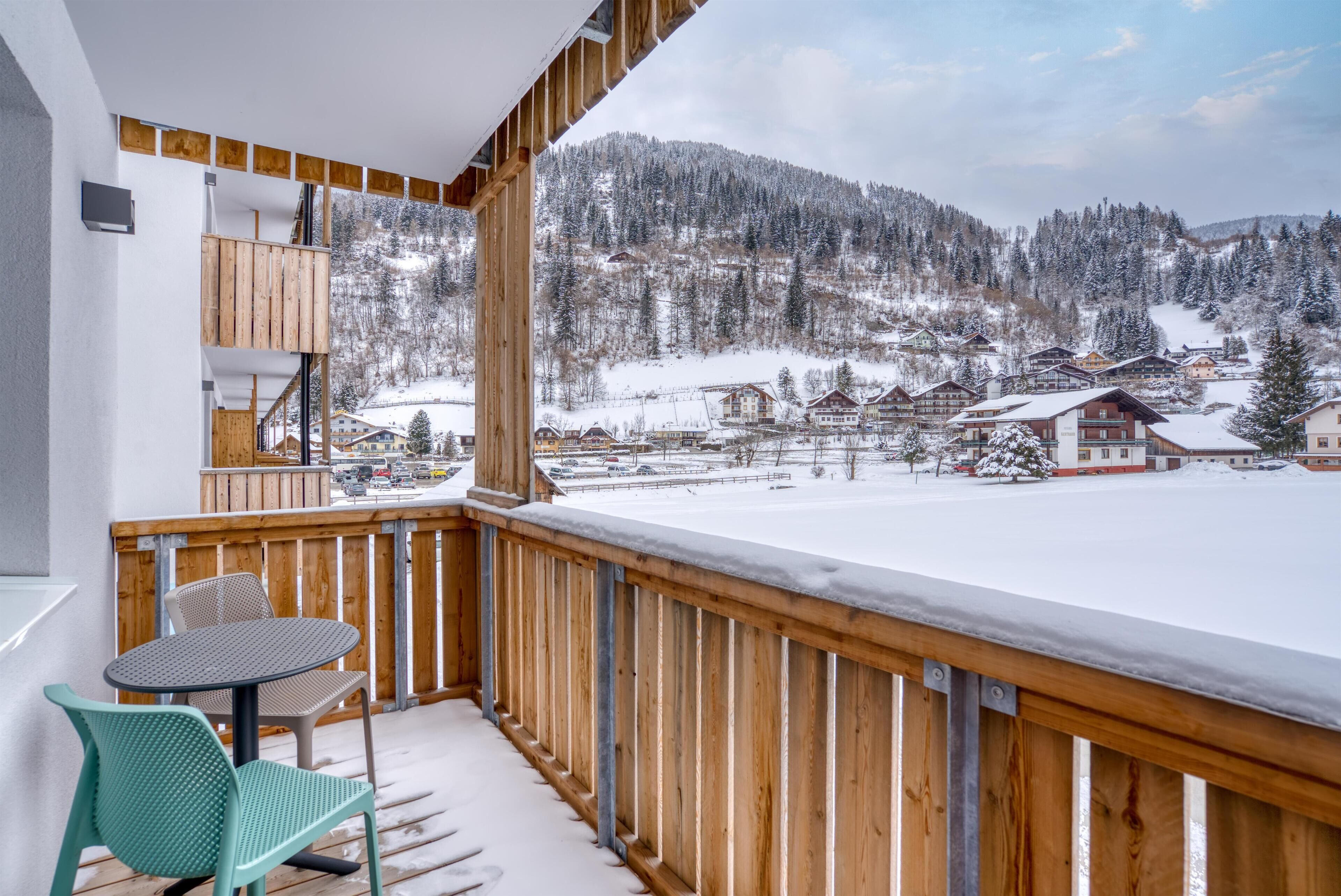 A balcony with a wooden railing overlooking a snow-covered village and mountains.