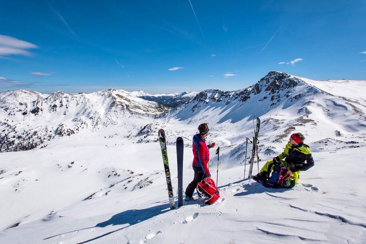 A snowy mountain landscape with two skiers, one standing and one sitting, with skis leaning against a pole.