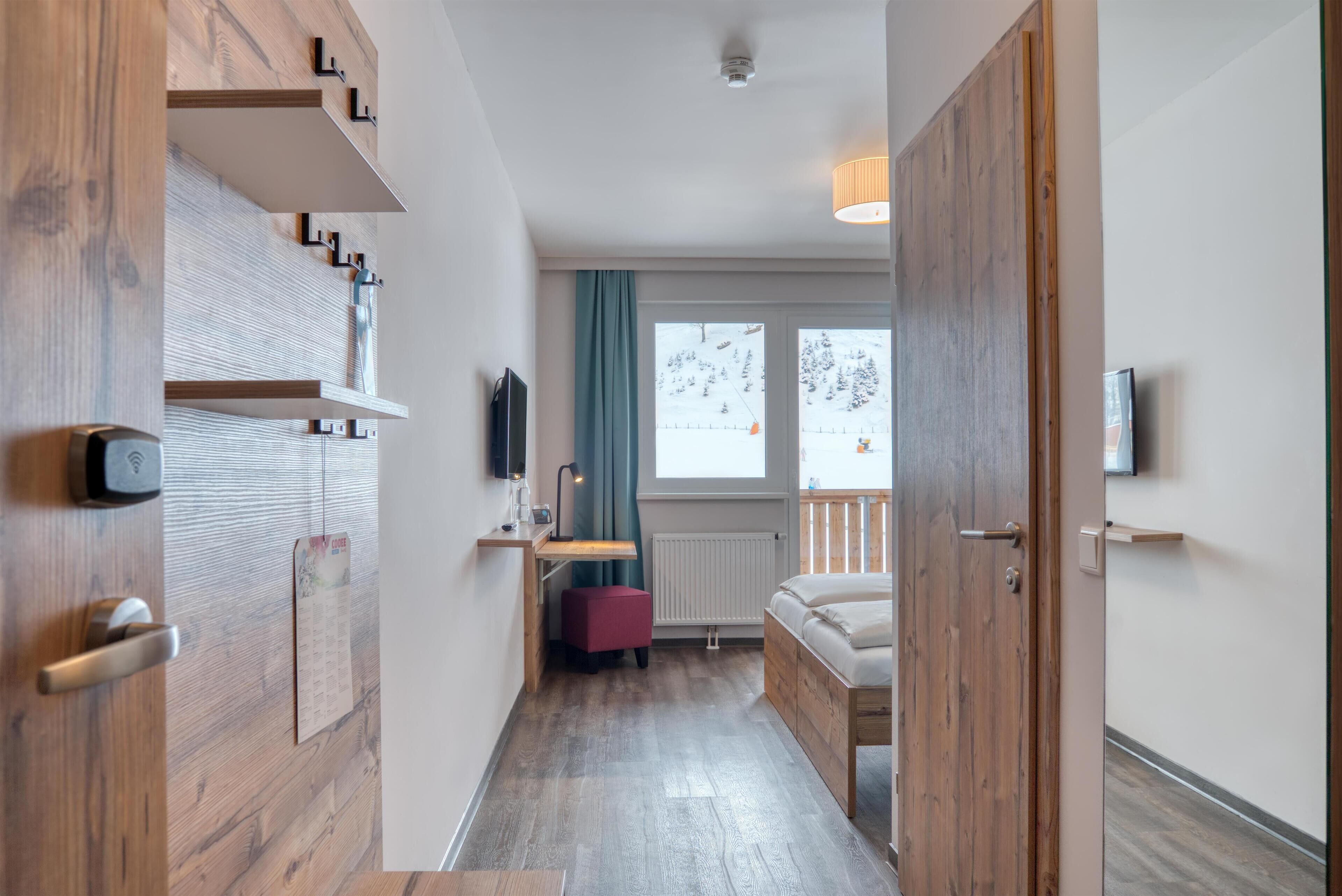 A modern hotel room with a wooden wardrobe, a desk with a chair, a bed, a TV, and a window with a view of a snowy landscape.