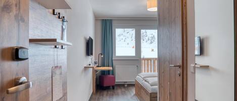 A modern hotel room with a wooden wardrobe, a desk with a chair, a bed, a TV, and a window with a view of a snowy landscape.