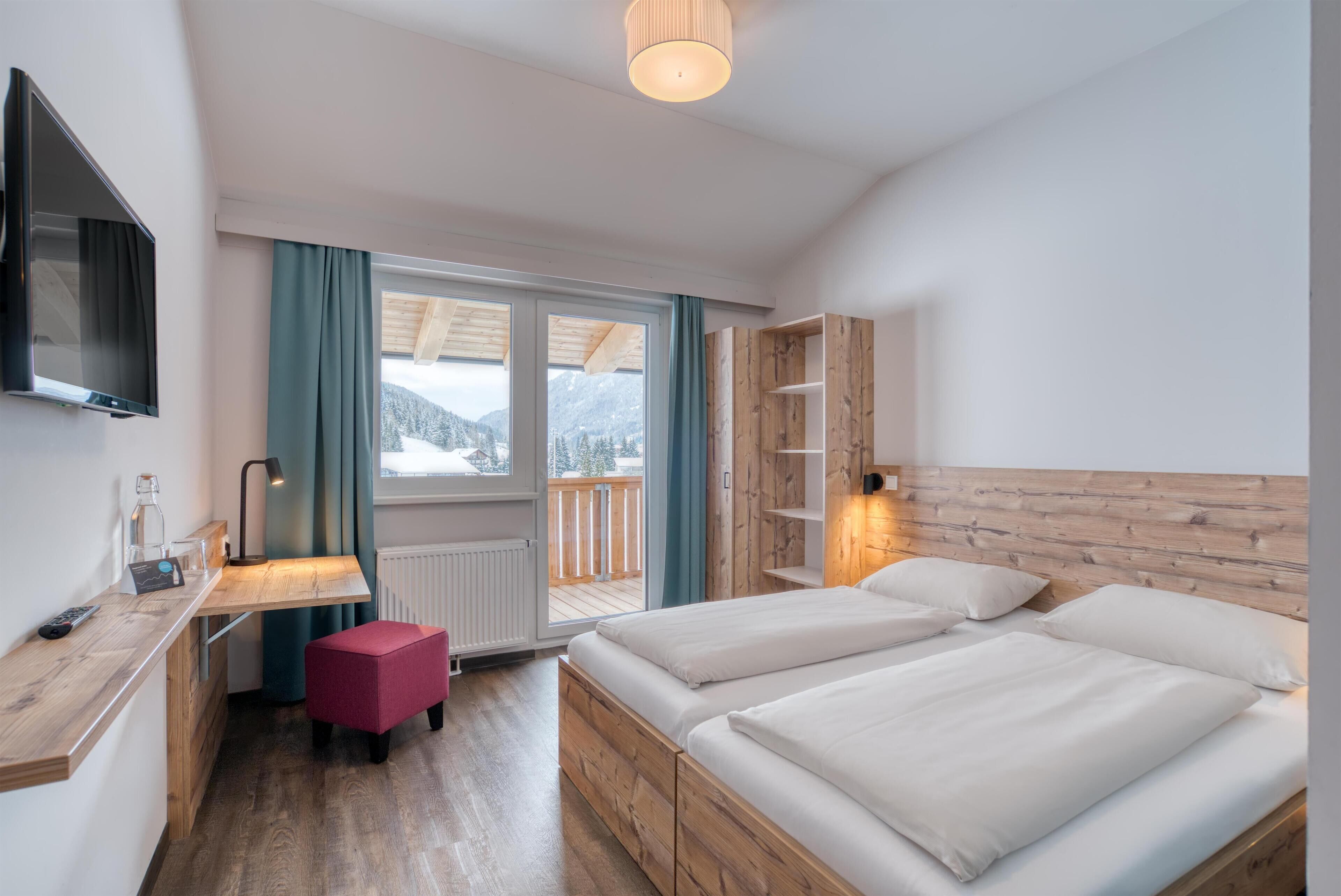 A modern hotel room with a wooden desk, a bed with two pillows, a red ottoman, a TV mounted on the wall, and a view of mountains through the window.