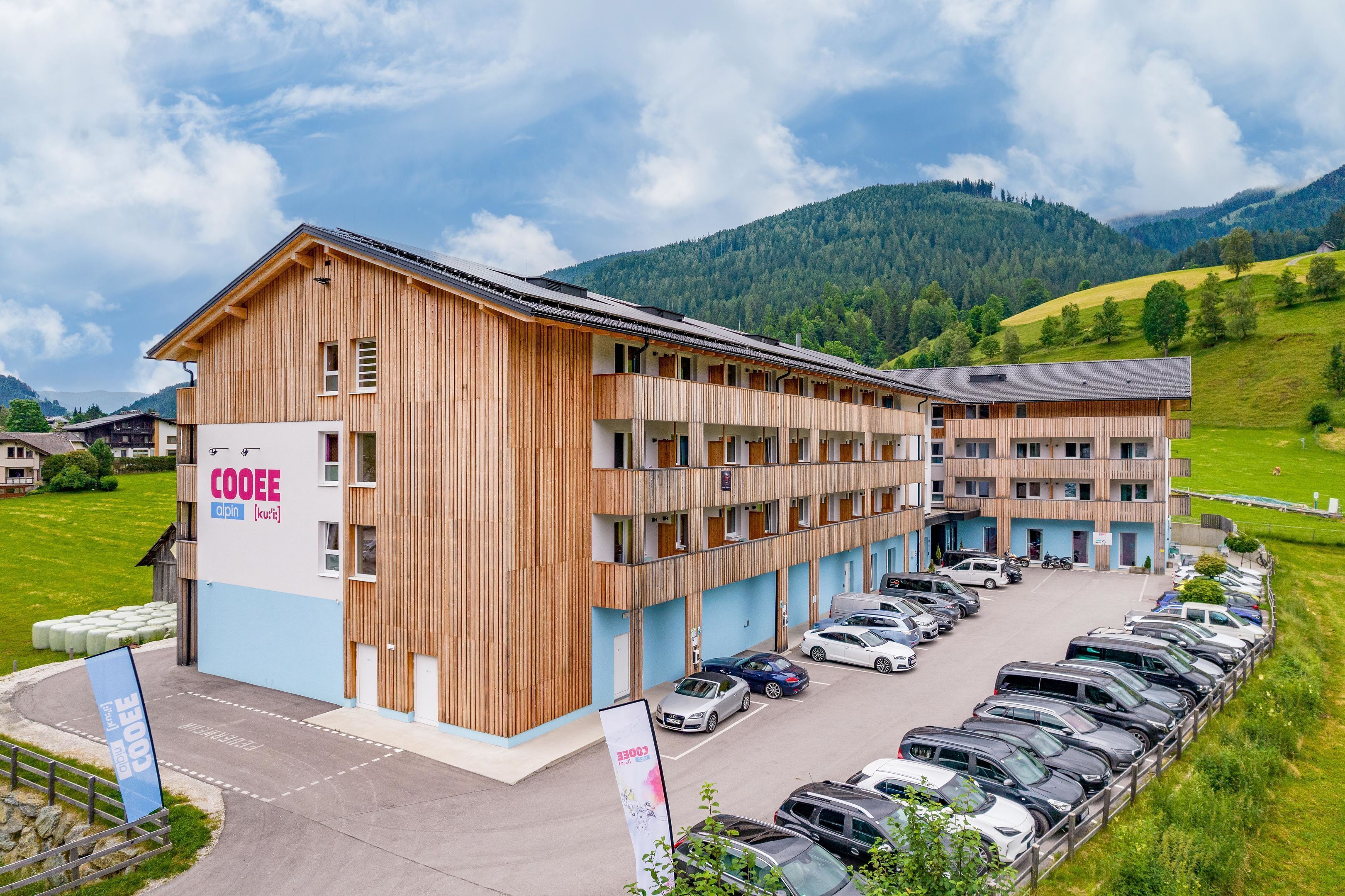 A multi-story hotel with a wooden facade, a parking lot, and a mountainous backdrop.