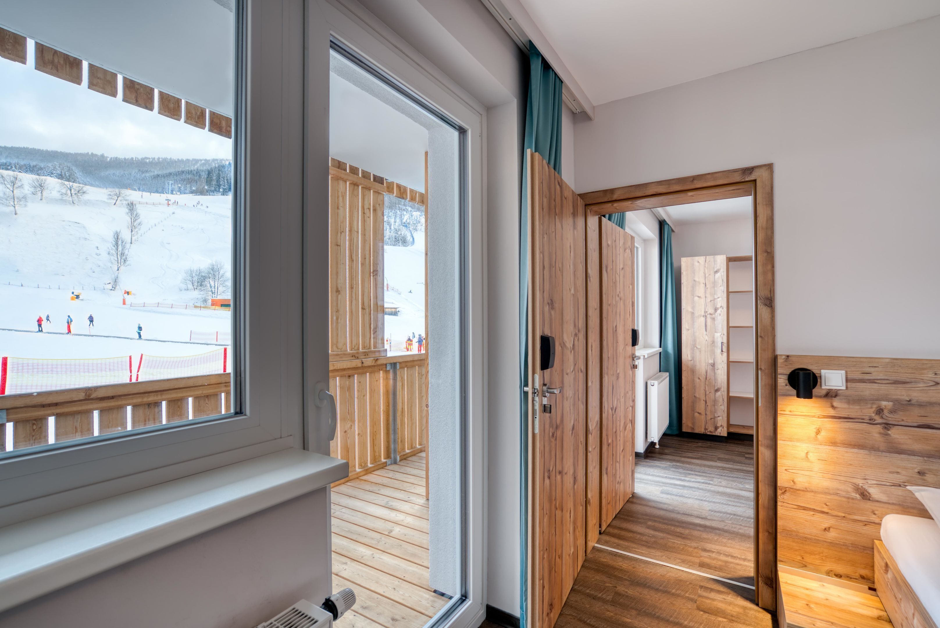Ein Zimmer mit Holzboden, einem Bett, einem Fenster mit Blick auf eine verschneite Landschaft und einer Holztür, die in ein weiteres Zimmer führt.