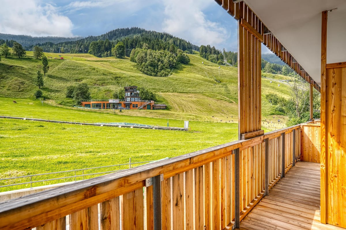 A wooden balcony overlooking a green field with a building in the distance.