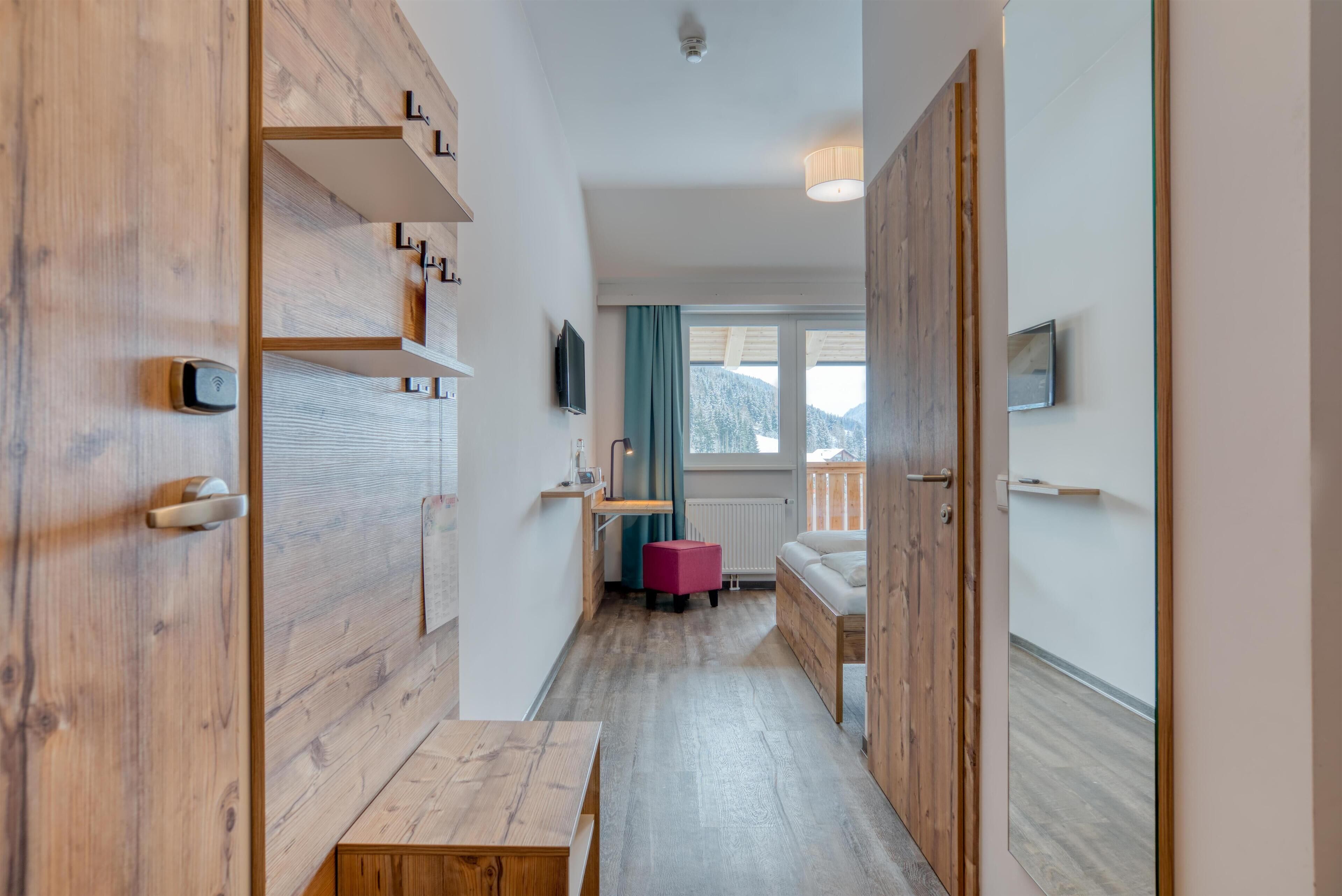 A narrow hallway with wooden flooring and walls, leading to a room with a window, a sofa, and a small table.
