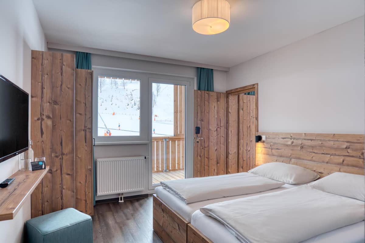 A bedroom with two beds, a TV, a wooden wardrobe, and a view of a snowy outdoor area.