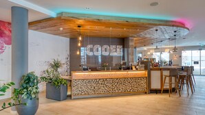 A modern reception area with a wooden ceiling, a counter with a stone pattern, and potted plants. - COOEE alpin Hotel Bad Kleinkirchheim (Bad Kleinkirchheim)