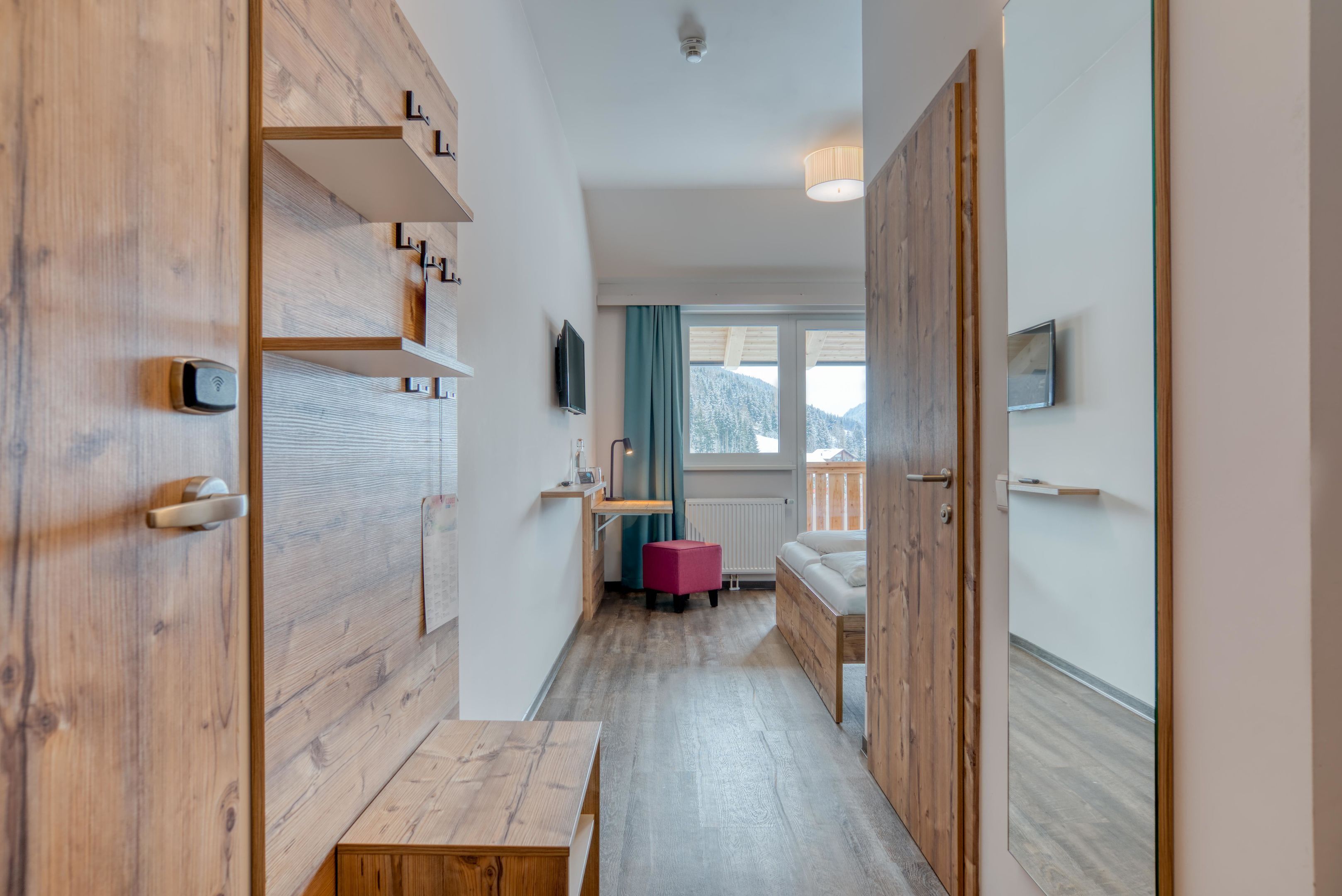 A narrow hallway with wooden flooring, a bench, a small table, a radiator, a window with a view of mountains, and a sofa in the background.