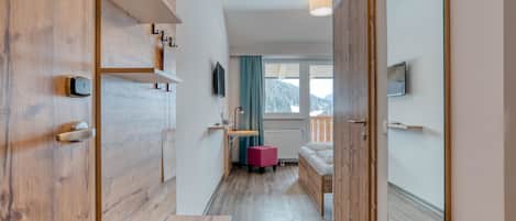 En smal gang med trægulv, en bænk, et lille bord, en radiator, et vindue der viser udsigt til bjerge, og en sofa i baggrunden.