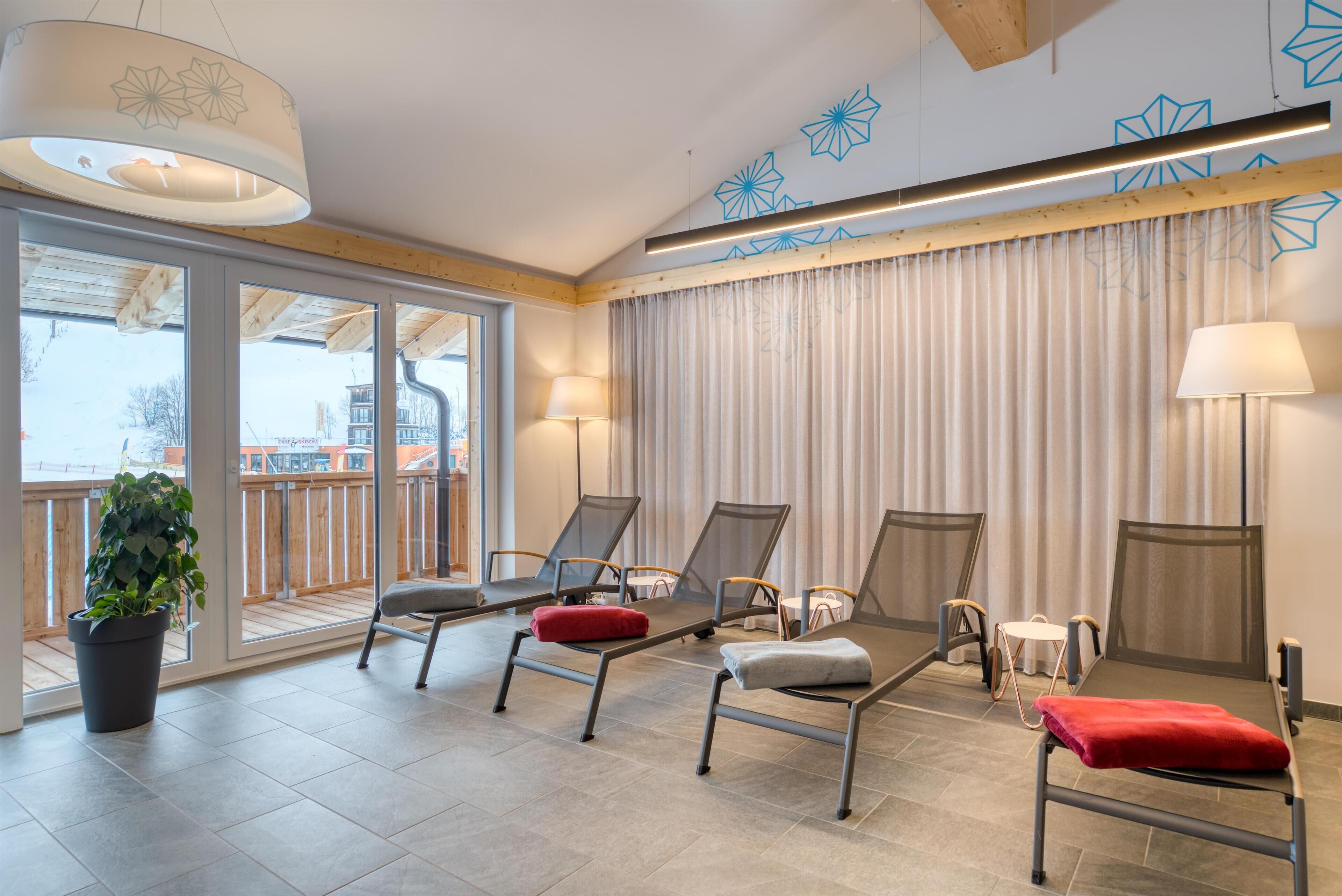 Un salón moderno con ventanas amplias, una planta en maceta y varios sillones con cojines rojos y grises.