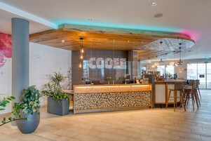 A reception desk with a wooden ceiling, a glass partition, and a sign that reads 'COOEE'. There are potted plants, a bar area with chairs, and a tiled floor.