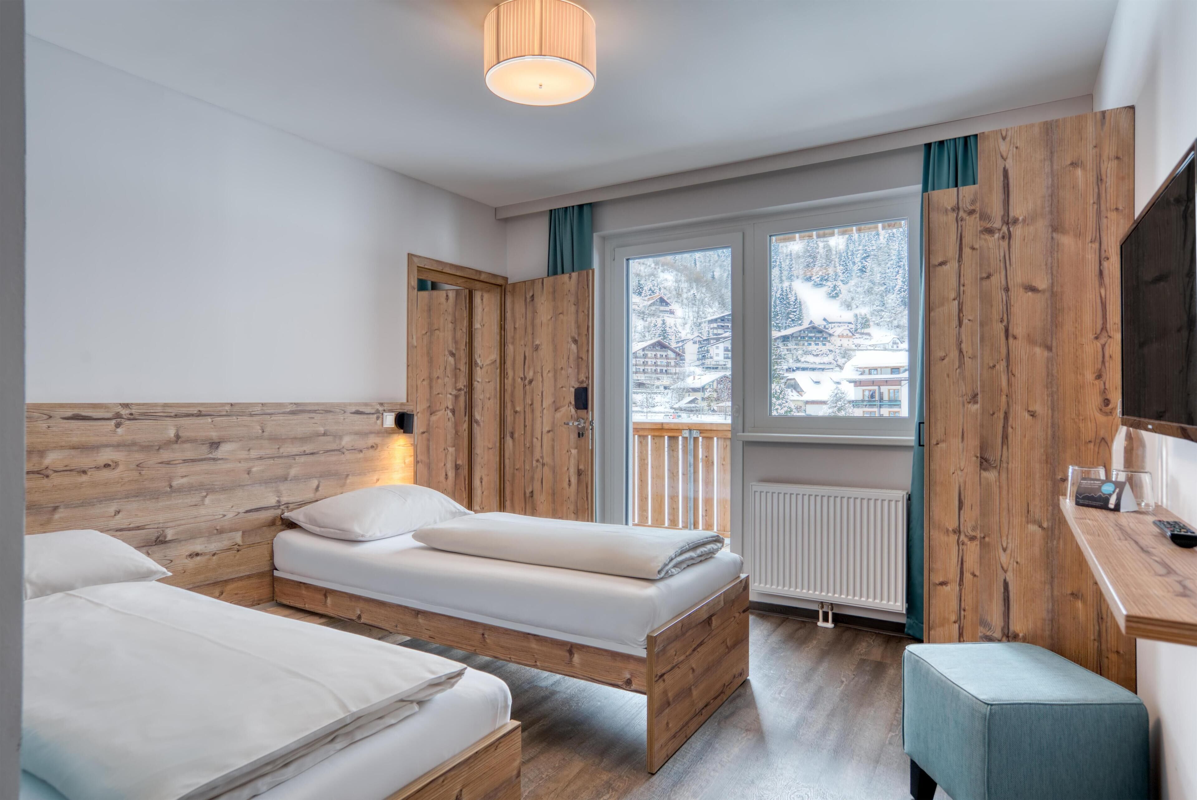Ein Zimmer mit zwei Betten, Holzwänden, einem Fenster mit Blick auf schneebedeckte Gebäude und einem Schreibtisch mit Stuhl.